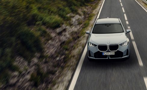  Gemini hat gesagt Dieses Bild zeigt einen modernen, hellgrauen BMW aus einer leicht erhöhten Frontalperspektive, während er eine asphaltierte Landstraße befährt. Die Umgebung ist von einer grünlich-braunen Hügellandschaft geprägt, wobei eine deutliche Bewegungsunschärfe an den Rändern der Aufnahme ein hohes Tempo und Dynamik suggeriert. Im Gegensatz zu den vorherigen Motiven verzichtet diese Variante komplett auf Textelemente oder grafische Rahmen, wodurch der Fokus allein auf dem Fahrzeug und der atmosphärischen Fahrtstrecke liegt.