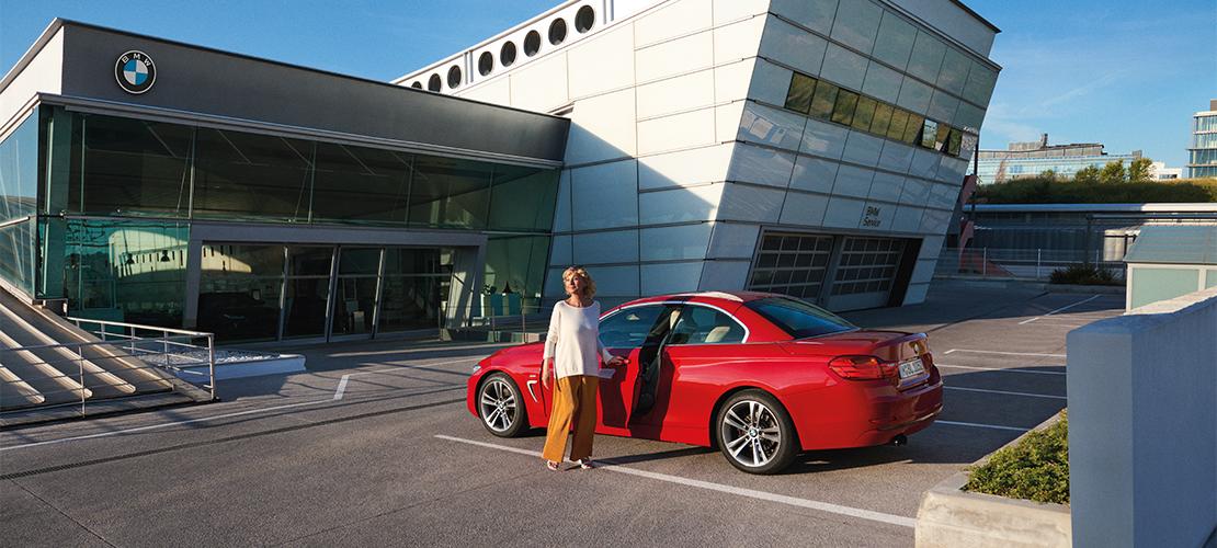 Eine Frau in einer hellen Jacke und orangefarbener Hose steht neben einem roten BMW Coupé mit geöffneter Fahrertür auf dem Parkplatz vor einem modernen BMW Autohaus oder Servicezentrum. Das Gebäude hat eine markante weiße Fassade mit Fensterreihen