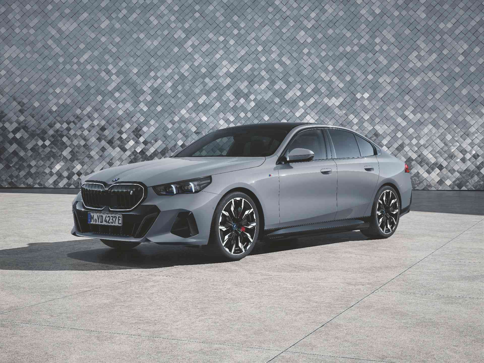 BMW i5 Limousine Ein silbergraues Auto steht auf einem hellgrauen gepflasterten Bereich vor einer Wand mit einem grauen, geometrischen Fliesenmuster. Das Auto hat schwarze Akzente und Felgen. Es ist leicht nach links gedreht, sodass die Vorderseite und die Fahrerseite zu sehen sind.