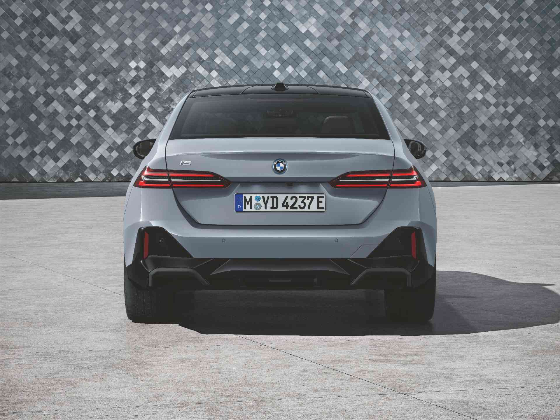 BMW i5 Limousine Ein silbergraues Auto steht auf einem hellgrauen gepflasterten Bereich vor einer Wand mit einem grauen, geometrischen Fliesenmuster. Das Auto hat schwarze Akzente und Felgen. Es ist leicht nach links gedreht, sodass die Vorderseite und die Fahrerseite zu sehen sind.