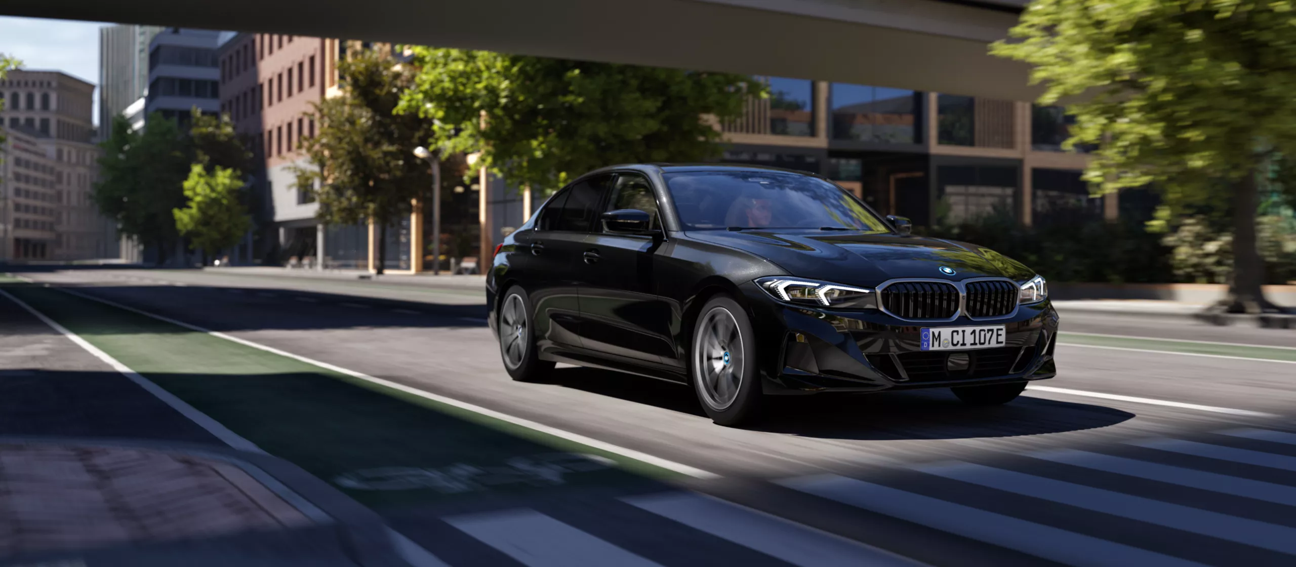 Das Bild zeigt eine schwarze BMW Limousine (vermutlich ein Modell der 3er-Reihe) in dynamischer Fahrt. Das Auto fährt auf einer breiten Stadtstraße mit einer grün markierten Spur (wahrscheinlich ein Radweg oder eine Busspur) und überquert gerade einen Zebrastreifen. Die Aufnahme ist aus einer niedrigen Perspektive und leicht von vorne gemacht, was dem Bild viel Geschwindigkeit und Bewegung verleiht. Im Hintergrund sind eine moderne Stadtkulisse mit mehrstöckigen Gebäuden aus Ziegeln und Glas sowie Bäumen entlang der Straße zu sehen. Die Beleuchtung ist hell und suggeriert Tageslicht. Das Kennzeichen des Wagens ist mit "M CI 107E" lesbar.