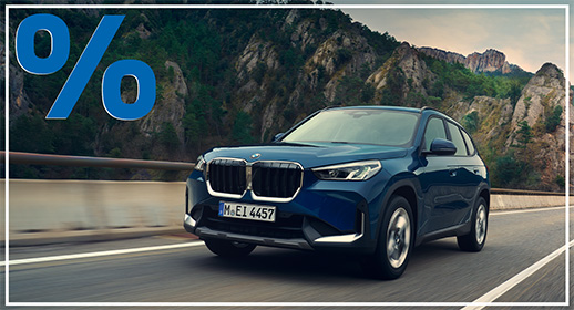 Das Bild zeigt einen dunkelblauen BMW X1, der auf einer Straße durch eine bergige Landschaft fährt. Auf der linken Seite befindet sich ein großes, rotes Prozentsymbol.