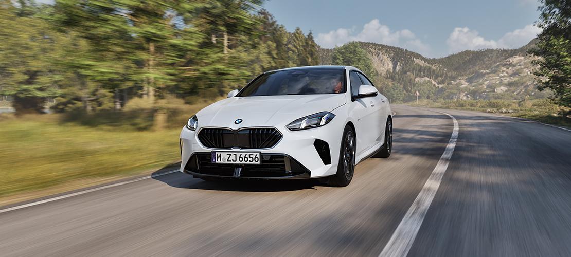 Es handelt sich um ein weißes BMW 2er Gran Coupé, das dynamisch auf einer kurvigen Landstraße unterwegs ist. Das Auto ist in der Mitte des Bildes von vorne zu sehen. Der Kühlergrill ist schwarz, und die LED-Scheinwerfer sind scharf gezeichnet. Im Hintergrund sind grüne Bäume und Berge unter einem hellen Himmel. Das Bild ist so aufgenommen, dass es Geschwindigkeit vermittelt, was durch die leichte Unschärfe des Hintergrundes noch verstärkt wird. Das Kennzeichen lautet "M ZJ 6656".