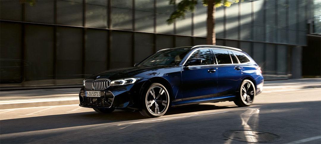 BMW 318i Touring Aktionsangebot Businesskunden Ein dunkelblauer BMW Kombi fährt auf einer Straße in einer städtischen Umgebung mit Gebäuden und einem Baum im Hintergrund, von der Seite betrachtet