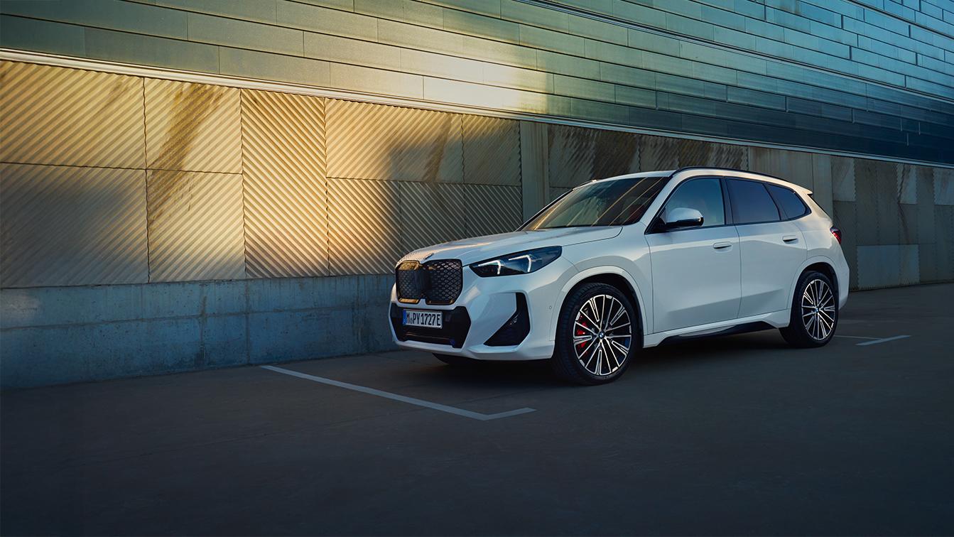 Weißer BMW iX1 in Front-Seitenansicht vor einer modernen Gebäudefassade bei warmem Sonnenlicht.