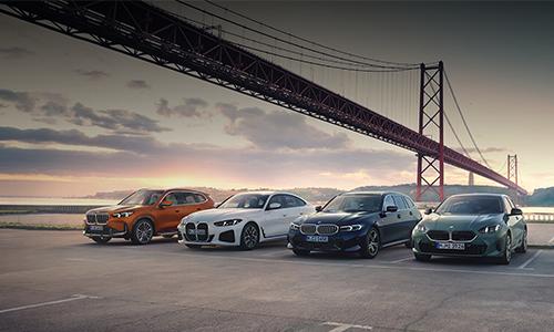 Vor der monumentalen Kulisse einer roten Hängebrücke präsentiert BMW vier aktuelle Modelle in der warmen Abendsonne. Auf einem weiten Parkplatz reihen sich ein orangefarbener X1, ein weißes 4er Gran Coupé, ein dunkelblauer 3er Touring und ein graugrüner 1er nebeneinander auf.  Die Szenerie am Wasser, die diagonale Stahlkonstruktion der Brücke und das goldene Licht verleihen der Aufnahme eine hochwertige, urbane Atmosphäre, die Vielseitigkeit und modernen Lifestyle ausstrahlt.