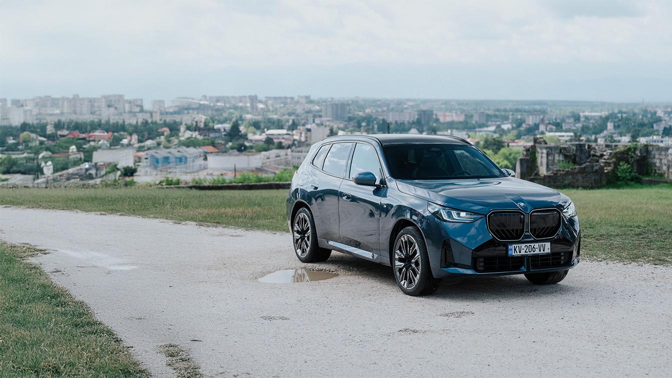 Ein graublauer BMW X3 steht auf einem hellen Schotterweg im Vordergrund, während sich im Hintergrund eine weitläufige Stadtlandschaft unter bewölktem Himmel erstreckt.