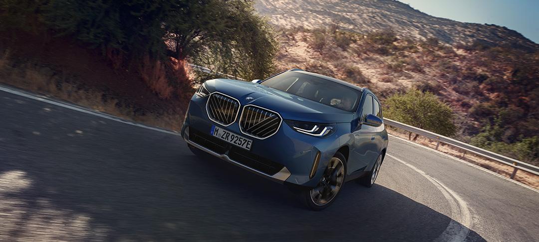 Das Bild zeigt einen modernen, dunkelblauen BMW X3 Plug-in-Hybriden in einer dynamischen Kurvenfahrt auf einer Serpentinenstraße. Durch die niedrige Kameraperspektive und die leichte Schräglage wirkt das SUV besonders kraftvoll und präsent. Markante Designelemente sind die neu gestaltete, große BMW-Niere mit diagonalen Streben und die flachen LED-Scheinwerfer. Die mediterran anmutende Hügellandschaft und das warme, goldene Licht der tiefstehenden Sonne verleihen der Szene eine hochwertige und sportliche Atmosphäre.