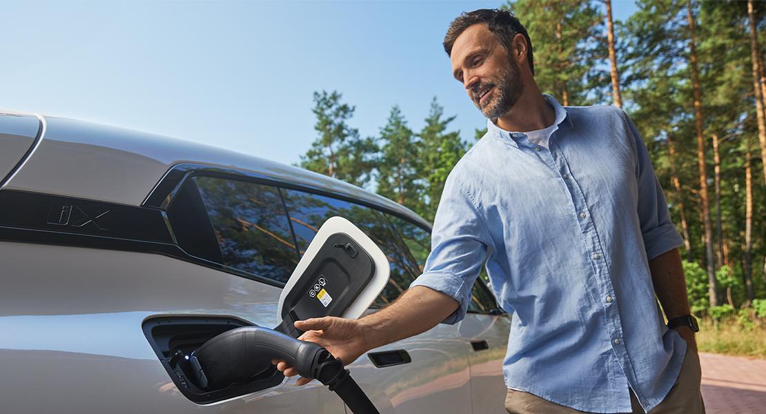 In einer hellen, freundlichen Außenaufnahme wird die unkomplizierte Handhabung moderner Elektromobilität thematisiert. Ein lächelnder Mann in einem legeren hellblauen Hemd steht entspannt neben seinem weißen Elektrofahrzeug, einem BMW iX, und führt gerade den Ladestecker in die vorgesehene Öffnung am Heck ein. Sein Blick ist zufrieden auf den Ladeanschluss gerichtet, was die intuitive Bedienung der Technik unterstreicht. Im Hintergrund ragen hohe Nadelbäume in den strahlend blauen Himmel, was dem technischen Vorgang eine natürliche und frische Note verleiht und die Mobilität als Teil eines aktiven Lebensstils darstellt.