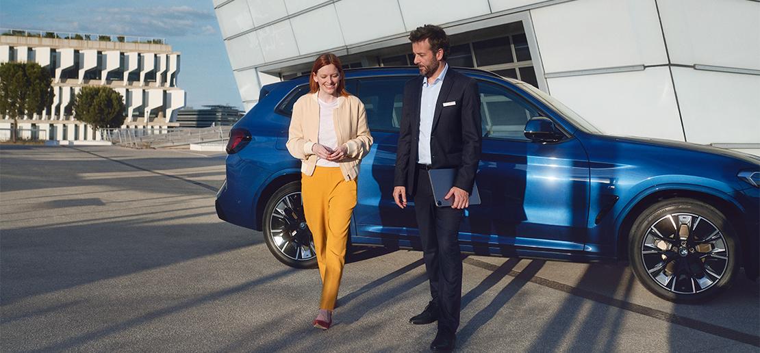 Eine lächelnde Frau in gelber Hose und beige Jacke und ein Mann im Anzug mit Tablet gehen nebeneinander vor einem blauen BMW SUV und moderner Architektur.