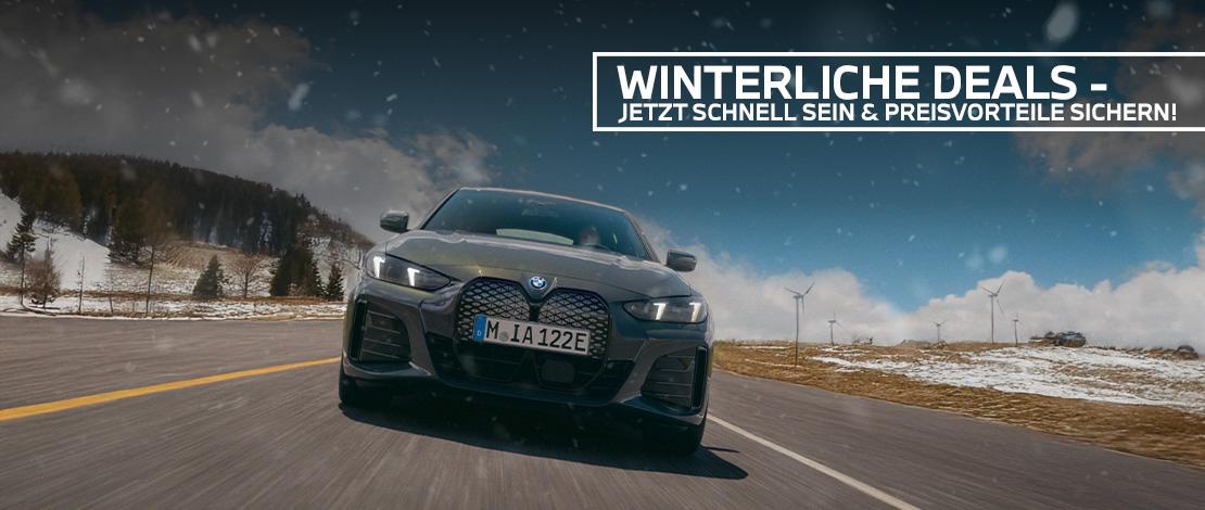 Ein dunkles, sportliches BMW 4er Coupé fährt frontal auf den Betrachter zu auf einer breiten Straße, die durch eine verschneite Landschaft führt. Im Hintergrund sind verschneite Bäume und Windräder unter einem blauen Himmel zu sehen. Ein weißer Textbanner über dem Auto lautet: "WINTERLICHE DEALS - JETZT SCHNELL SEIN & PREISVORTEILE SICHERN!". Das Bild vermittelt Geschwindigkeit und eine winterliche Verkaufsaktion.