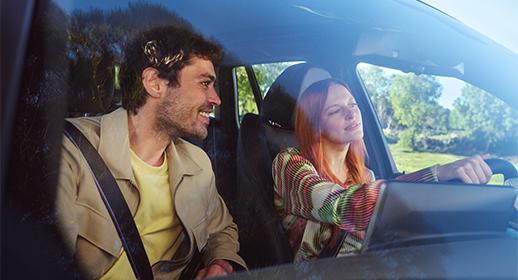 Das Bild zeigt ein entspanntes Paar während einer Autofahrt ins Grüne. Ein bärtiger Mann auf dem Beifahrersitz blickt lächelnd zur Seite, während eine rothaarige Frau am Steuer konzentriert nach vorne schaut. Das helle Sonnenlicht und die Bäume im Hintergrund verleihen der Szene eine freundliche, sommerliche Atmosphäre.
