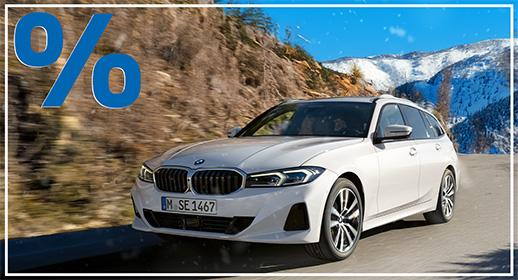 Ein weißer BMW 3er Touring fährt auf einer Bergstraße. Im Hintergrund erheben sich schneebedeckte Berge und bewaldete Hänge unter blauem Himmel. Das Auto ist frontal von schräg rechts zu sehen. Das blaue Prozentzeichen (%) füllt die obere linke Ecke und signalisieren einen Rabatt oder eine spezielle Finanzierung.
