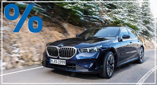 Eine dunkelblaue BMW 5er Limousine ist frontal leicht von links auf einer kurvigen Straße zu sehen. Die Umgebung ist ein verschneiter Wald mit Tannenbäumen. Die Szene wirkt dynamisch mit leicht fallendem Schnee. Das blaue Prozentzeichen (%) füllt die obere linke Ecke und signalisieren einen Rabatt oder eine spezielle Finanzierung.