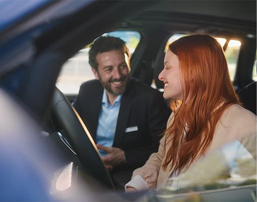 Ein Mann auf dem Beifahrersitz blickt die lächelnde Fahrerin an. Sonnenlicht fällt durch das Fenster.