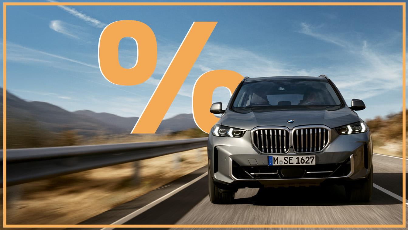 Das Bild zeigt einen grauen BMW X5, der auf einer Straße fährt. Im Hintergrund sind Berge zu sehen. Auf der linken Seite des Bildes befindet sich ein großes, oranges Prozentsymbol. Ein orangefarbener Rahmen umgibt das gesamte Bild.