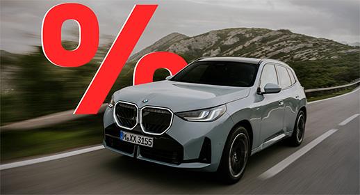  Das Bild zeigt einen hellgrauen, viertürigen BMW X3, der auf einer Landstraße fährt. Das Auto ist schräg von vorne zu sehen. Im Vordergrund ist ein großes, rotes Prozentzeichen (%) platziert. Der Hintergrund zeigt eine bergige Landschaft unter einem bewölkten Himmel. Das Kennzeichen "M XX 3155" ist auf der vorderen Stoßstange zu sehen.