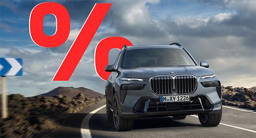 Dieses Bild zeigt ein großes, graues SUV, den BMW X7, auf einer kurvigen Straße in einer bergigen Landschaft. Das Auto fährt direkt auf den Betrachter zu. Oben links im Bild ist ein sehr großes, rotes Prozentzeichen (%) platziert. Das Kennzeichen des Autos lautet "M XY 1226".