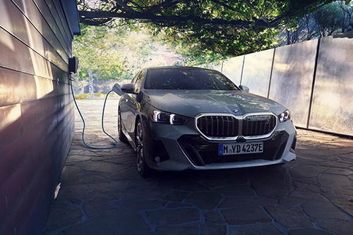 Dieses Bild zeigt einen hellgrauen Plug-in-Hybrid oder ein Elektrofahrzeug beim Laden unter einem Vordach. Das Auto steht in einer leicht gedimmten Einfahrt. Links an einer dunkelbraun verkleideten Hauswand ist eine weiße Wallbox installiert. Ein hellblaues Kabel führt von der Wallbox in den Ladeanschluss des Fahrzeugs. Die Decke des Carports ist dicht mit Laub und Ästen bewachsen, was eine natürliche, schattige Atmosphäre schafft, typisch für eine private Ladesituation in den Abendstunden.